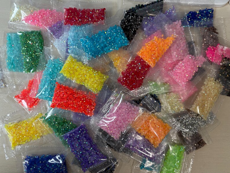 May include: A collection of small, clear plastic bags filled with multi-colored, faceted resin rhinestones. The assortment includes vibrant shades of red, orange, yellow, green, blue, purple, pink, black, and silver, all featuring a reflective finish.