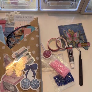May include: A craft kit containing a brown polka dot envelope filled with stationery, a butterfly sticker, a disco ball cherry sticker, two rolls of pink washi tape, a tube of B-7000 glue, black tweezers, and two small bags of pink and blue rhinestones.