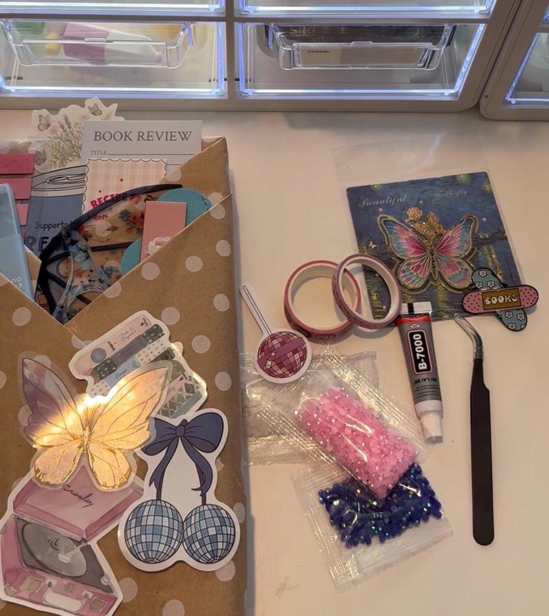 May include: A craft kit containing a brown polka dot envelope filled with stationery, a butterfly sticker, a disco ball cherry sticker, two rolls of pink washi tape, a tube of B-7000 glue, black tweezers, and two small bags of pink and blue rhinestones.