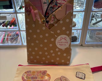 May include: A tan envelope with white polka dots containing stationery items, including a book review card, a soda can-shaped notepad, and clips. Below is a cream-colored canvas pouch with a pink base, a floral paperclip, two rolls of washi tape, and a book-shaped pin.