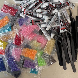 May include: A collection of craft supplies including numerous small plastic bags filled with colorful faceted rhinestones, several tubes of B-7000 adhesive, multiple black metal tweezers, and a row of white pencils with light-colored tips.