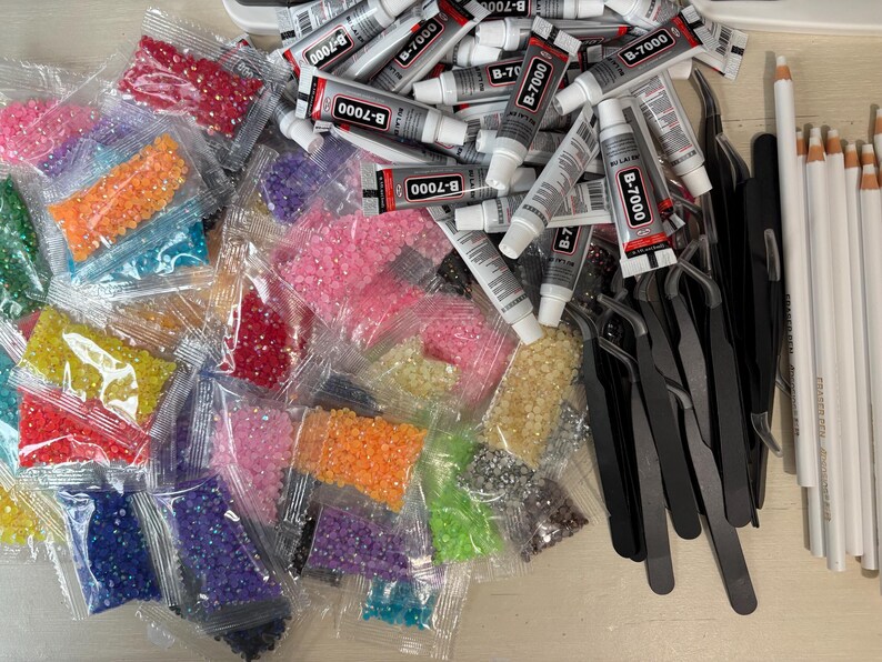May include: A collection of craft supplies including numerous small plastic bags filled with colorful faceted rhinestones, several tubes of B-7000 adhesive, multiple black metal tweezers, and a row of white pencils with light-colored tips.