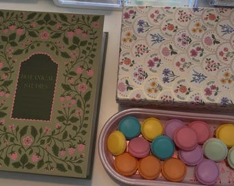 May include: A green book titled Botanical Studies with a strawberry vine pattern, a rectangular box with a floral scalloped print, and a light pink oval tray containing an assortment of colorful round macarons in yellow, blue, pink, and orange.