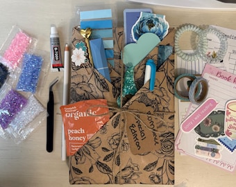 May include: A craft kit featuring a brown floral paper envelope tied with twine, containing a blue pen, a heart-topped pin, and a peach honey packet. Accessories include five small bags of colorful rhinestones, tweezers, glue, two hair coils, washi tape, stickers, and a reading log.