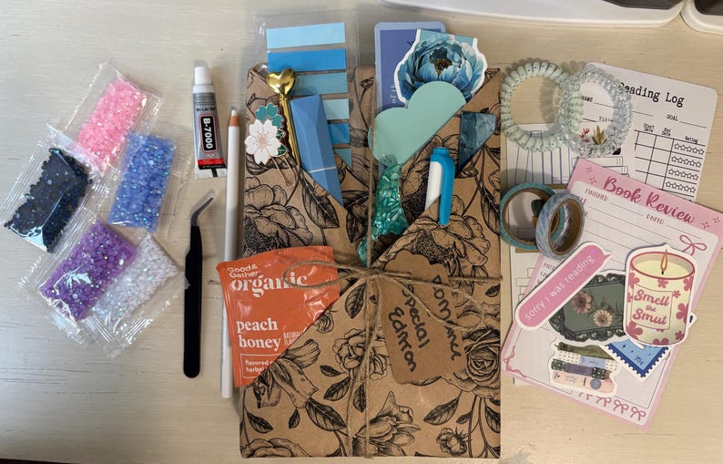 May include: A craft kit featuring a brown floral paper envelope tied with twine, containing a blue pen, a heart-topped pin, and a peach honey packet. Accessories include five small bags of colorful rhinestones, tweezers, glue, two hair coils, washi tape, stickers, and a reading log.