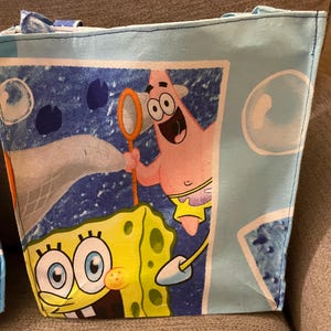 May include: Light blue tote bag featuring SpongeBob SquarePants and Patrick Star from the animated series. The bag has a rectangular shape with a light blue background and a cartoon graphic. The bag has two handles.