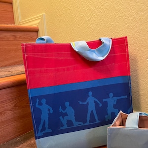 Puede incluir: Dos bolsas de tela azules y rojas con un dise&ntilde;o de rayas azules y blancas. La bolsa m&aacute;s grande tiene una silueta de cinco figuras en la raya. La bolsa m&aacute;s peque&ntilde;a tiene el texto "To Infinity and Beyond" en la parte delantera.