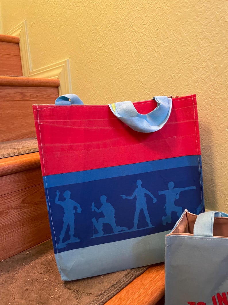 Puede incluir: Dos bolsas de tela azules y rojas con un dise&ntilde;o de rayas azules y blancas. La bolsa m&aacute;s grande tiene una silueta de cinco figuras en la raya. La bolsa m&aacute;s peque&ntilde;a tiene el texto "To Infinity and Beyond" en la parte delantera.