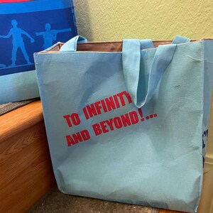 Puede incluir: Una bolsa de tela azul claro con asas marrones. La bolsa tiene el texto "To Infinity and Beyond!" impreso en rojo en la parte delantera.