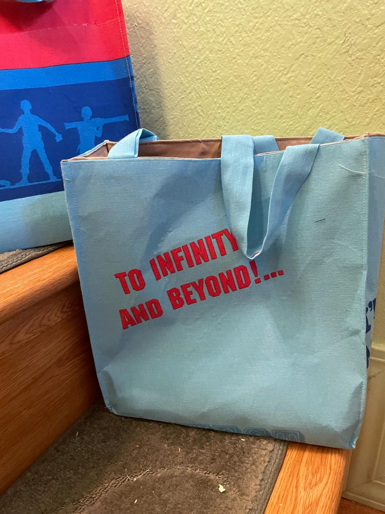 Puede incluir: Una bolsa de tela azul claro con asas marrones. La bolsa tiene el texto "To Infinity and Beyond!" impreso en rojo en la parte delantera.