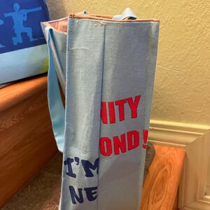 Puede incluir: Una bolsa de tela azul claro con asas marrones. La bolsa tiene el texto "Unity Beyond! I'm One" impreso en rojo y azul.