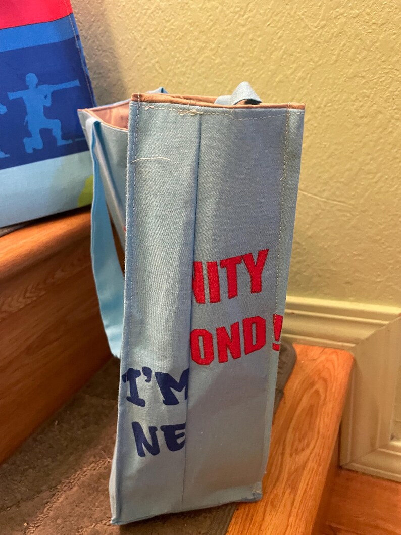 Puede incluir: Una bolsa de tela azul claro con asas marrones. La bolsa tiene el texto "Unity Beyond! I'm One" impreso en rojo y azul.