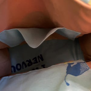 Puede incluir: Una bolsa de tela azul y rosa con un bolsillo en el interior. La bolsa tiene un forro de tela azul y blanco con el texto "No estoy nervioso" impreso en &eacute;l. La bolsa tambi&eacute;n tiene un gr&aacute;fico azul y amarillo de un animal estilizado.