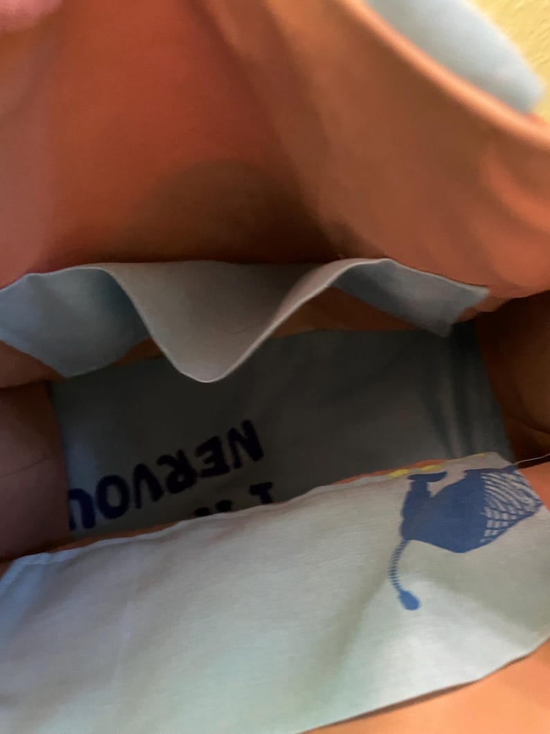 Puede incluir: Una bolsa de tela azul y rosa con un bolsillo en el interior. La bolsa tiene un forro de tela azul y blanco con el texto "No estoy nervioso" impreso en &eacute;l. La bolsa tambi&eacute;n tiene un gr&aacute;fico azul y amarillo de un animal estilizado.