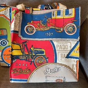 May include: Two tote bags with a vintage car theme. The bags feature colorful illustrations of antique cars, license plates, and the years 1903 and 1909. The bags have beige handles and are made of a sturdy fabric.