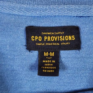 May include: A close-up of a light blue shirt collar with a black label. The label displays "SHIPMEN SUPPLY CPO PROVISIONS SIMPLE PRACTICAL UTILITY" in gold text. Below, it reads "M-M" and "MADE IN INDIA FABRIQUE EN INDE".