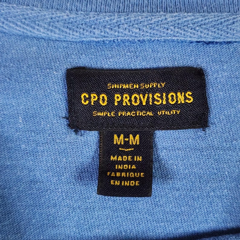 May include: A close-up of a light blue shirt collar with a black label. The label displays "SHIPMEN SUPPLY CPO PROVISIONS SIMPLE PRACTICAL UTILITY" in gold text. Below, it reads "M-M" and "MADE IN INDIA FABRIQUE EN INDE".