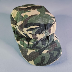May include: A military-style patrol cap featuring a green, brown, and black camouflage pattern. The hat is constructed from a grid-textured fabric and includes a flat, rounded brim and a structured crown with a band around the base.