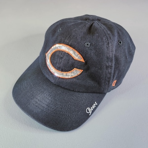 May include: A navy blue baseball cap featuring a large letter C on the front panel, outlined in orange thread and filled with silver sequins. The word Bears is embroidered in white script on the left side of the curved brim, with a small orange tag.