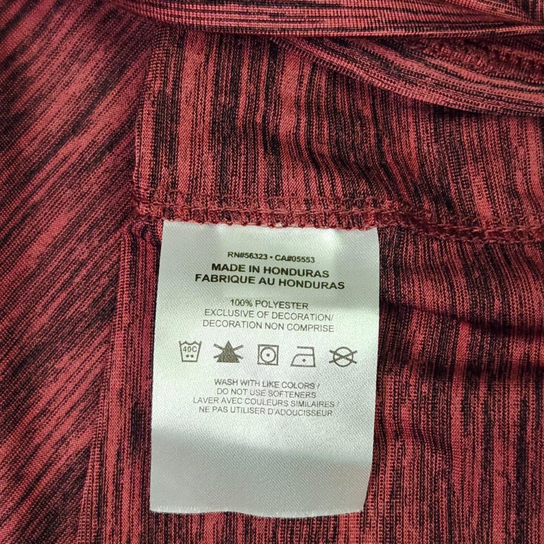 Puede incluir: Primer plano de una tela estampada en rojo y negro con una etiqueta blanca. La etiqueta incluye el texto: "MADE IN HONDURAS", "100% POLYESTER" e instrucciones de lavado. La tela tiene un aspecto texturizado.