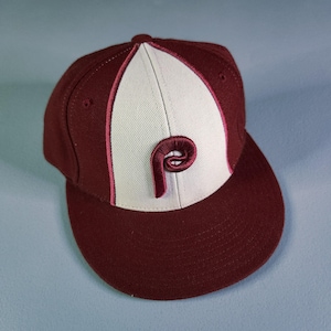 May include: A maroon and off-white baseball cap featuring a structured crown with a flat brim. The front panel is off-white with a maroon embroidered letter P in a cursive script style. The cap includes maroon side panels and maroon piping.