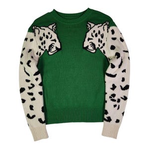 May include: A long-sleeve crewneck jumper featuring a solid green torso and white sleeves with a black leopard spot pattern. The upper chest area displays two white leopard head graphics with black facial details, positioned symmetrically.