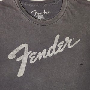 Puede incluir: Camiseta gris oscuro con la palabra "Fender" en letras blancas grandes y desgastadas en la parte delantera. La camiseta tiene cuello redondo y una peque&ntilde;a etiqueta con texto cerca del cuello.