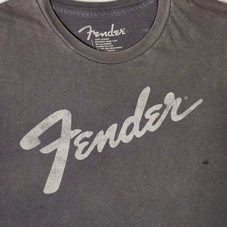 Puede incluir: Camiseta gris oscuro con la palabra "Fender" en letras blancas grandes y desgastadas en la parte delantera. La camiseta tiene cuello redondo y una peque&ntilde;a etiqueta con texto cerca del cuello.