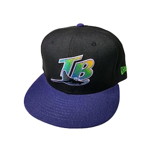 Puede incluir: Gorra de béisbol negra con visera morada. La gorra presenta las letras "TB" en un degradado de verde, amarillo y azul, con un diseño estilizado de manta raya. Un logotipo verde de New Era está en el lateral.