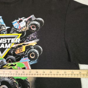 Puede incluir: Camiseta negra con un gr&aacute;fico colorido de monster trucks en acci&oacute;n. El dise&ntilde;o incluye el texto "MONSTER JAM" en negrita, junto con otros textos y logotipos. La imagen muestra varios dise&ntilde;os de monster trucks.