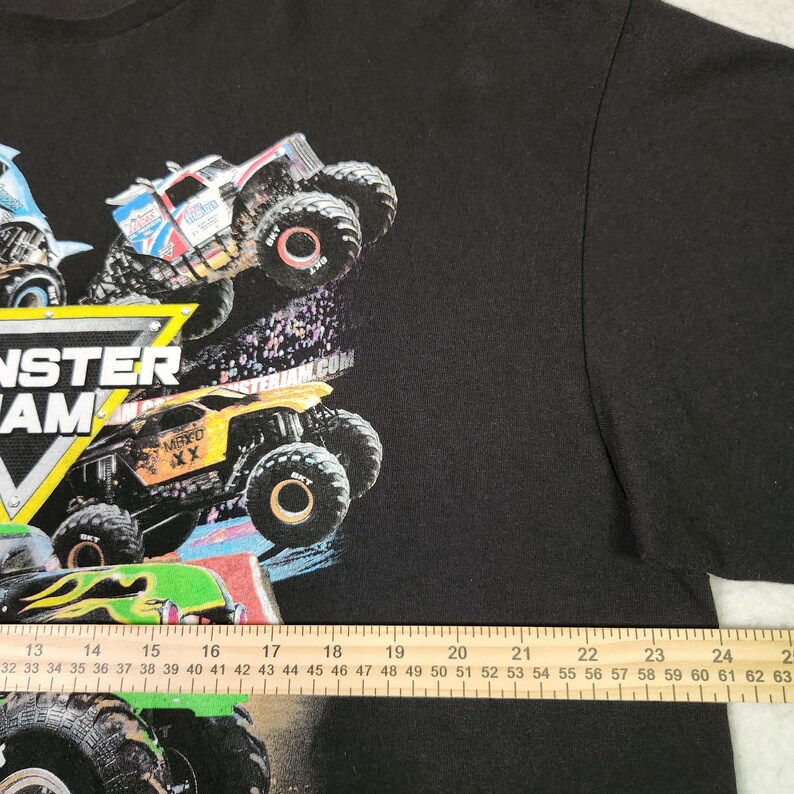Puede incluir: Camiseta negra con un gr&aacute;fico colorido de monster trucks en acci&oacute;n. El dise&ntilde;o incluye el texto "MONSTER JAM" en negrita, junto con otros textos y logotipos. La imagen muestra varios dise&ntilde;os de monster trucks.
