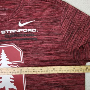Puede incluir: Una camiseta Nike Stanford Cardinal burdeos y negra. La camiseta presenta el logotipo de Stanford y la palabra "STANFORD" en blanco, con un logotipo de Nike blanco. Una regla se coloca en la parte inferior de la camisa, midiendo hasta 64 cm.