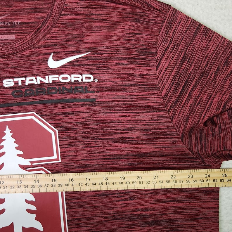 Puede incluir: Una camiseta Nike Stanford Cardinal burdeos y negra. La camiseta presenta el logotipo de Stanford y la palabra "STANFORD" en blanco, con un logotipo de Nike blanco. Una regla se coloca en la parte inferior de la camisa, midiendo hasta 64 cm.