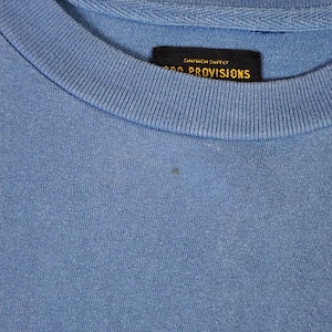 May include: A close-up shot of a light blue crew neck t-shirt. A black tag with gold lettering is sewn into the collar, displaying the text "SHIPMEN SUPPLY" and "PROVISIONS". The fabric has a slightly textured appearance.