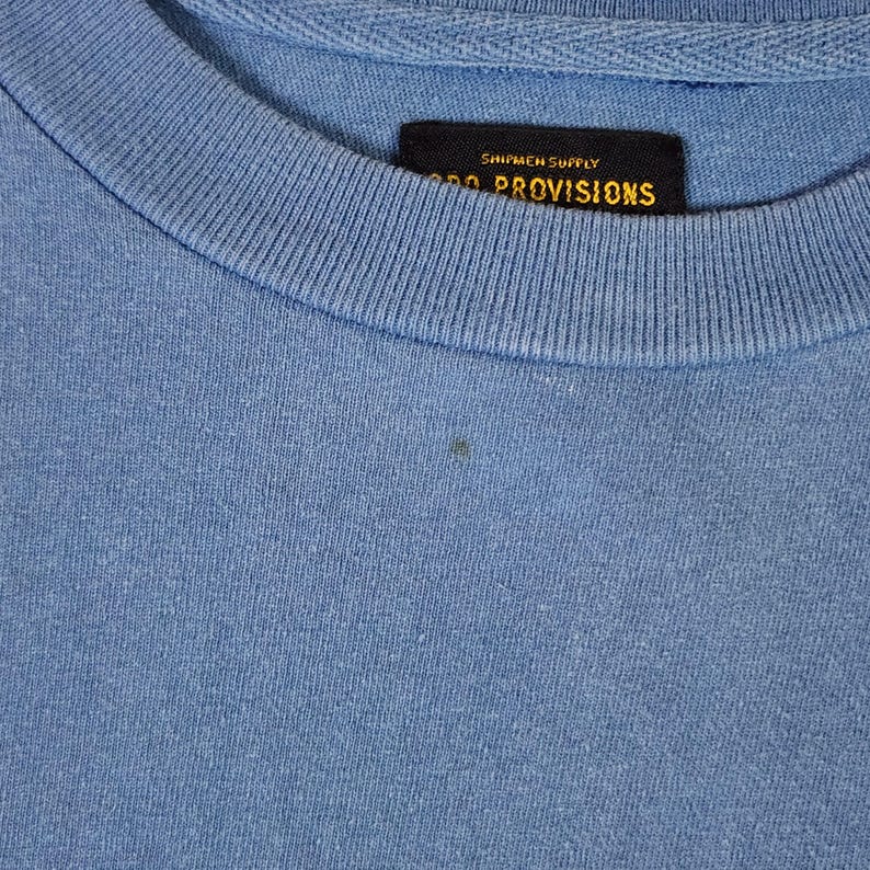May include: A close-up shot of a light blue crew neck t-shirt. A black tag with gold lettering is sewn into the collar, displaying the text "SHIPMEN SUPPLY" and "PROVISIONS". The fabric has a slightly textured appearance.
