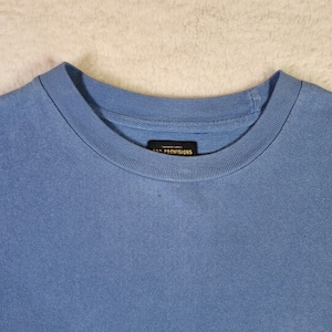 May include: A pale blue crew neck t-shirt. The shirt features a simple design with a ribbed neckline and a small rectangular label near the collar that reads "PROVISIONS". The material looks soft and comfortable.