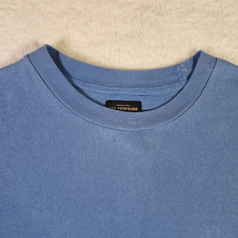 May include: A pale blue crew neck t-shirt. The shirt features a simple design with a ribbed neckline and a small rectangular label near the collar that reads "PROVISIONS". The material looks soft and comfortable.