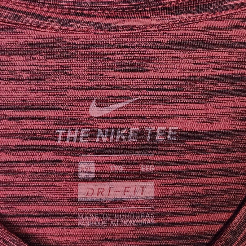 Puede incluir: Primer plano de una camiseta deportiva roja y negra con el logo de Nike y el texto "THE NIKE TEE". La camiseta tiene un patr&oacute;n texturizado y las palabras "DRI-FIT" son visibles.
