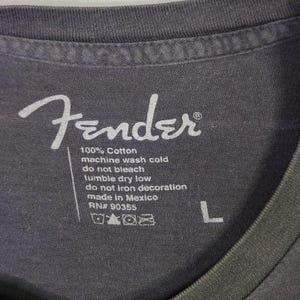 Puede incluir: Primer plano de una etiqueta de camiseta gris con el logotipo blanco de Fender y las instrucciones de cuidado. La etiqueta dice "100% Algod&oacute;n, lavar a m&aacute;quina en fr&iacute;o, no usar lej&iacute;a, secar en secadora a baja temperatura, no planchar la decoraci&oacute;n, hecho en M&eacute;xico, RN# 90355, L."