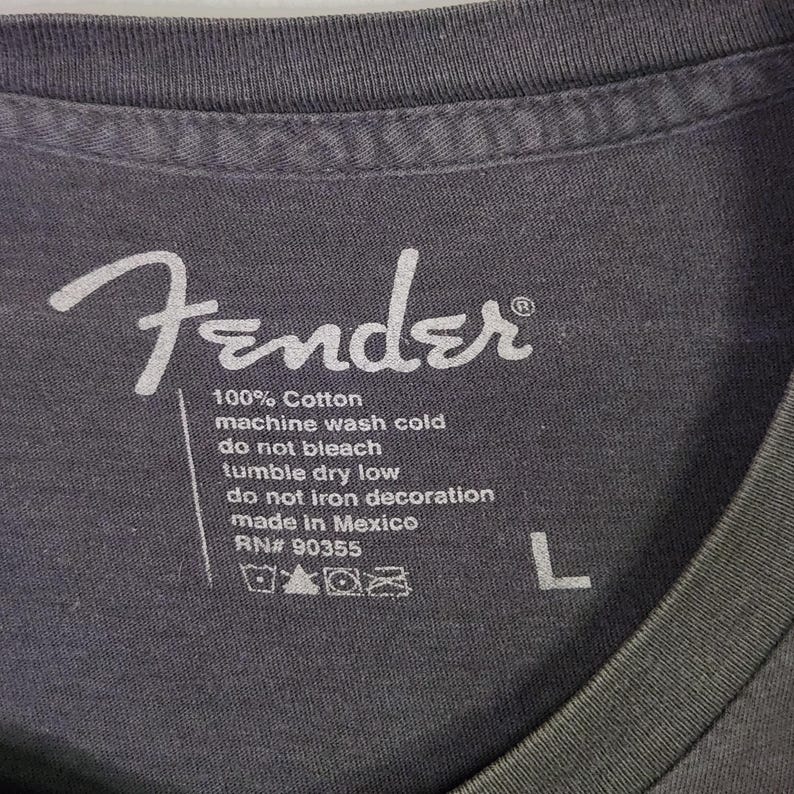 Puede incluir: Primer plano de una etiqueta de camiseta gris con el logotipo blanco de Fender y las instrucciones de cuidado. La etiqueta dice "100% Algod&oacute;n, lavar a m&aacute;quina en fr&iacute;o, no usar lej&iacute;a, secar en secadora a baja temperatura, no planchar la decoraci&oacute;n, hecho en M&eacute;xico, RN# 90355, L."