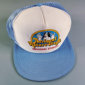 Puede incluir: Una gorra de camionero azul claro y blanca con un gráfico de una cacatúa, un perro y un gato dentro de un óvalo amarillo. El texto dice Animal Actors Stage y UNIVERSAL STUDIOS en rojo. La gorra tiene una parte trasera de malla y visera azul.