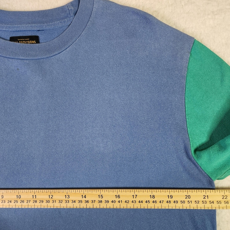 May include: A blue crew neck t-shirt with a green sleeve. The shirt has the word "PROVISIONS" on the collar. The shirt is measured with a ruler in centimetres.