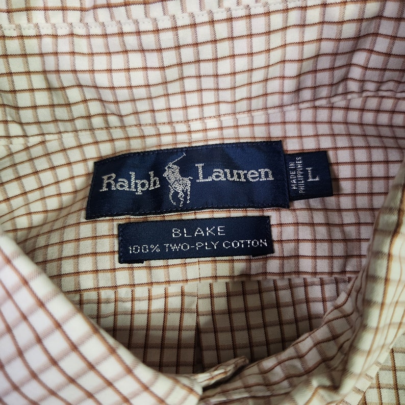 May include: A brown and white plaid shirt with a blue label that reads "Ralph Lauren" and "Blake 100% Two-Ply Cotton". The label also includes a small polo player logo and "Made in Philippines" in smaller text.