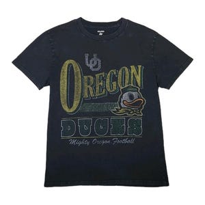 Puede incluir: Camiseta negra con la inscripción "Oregon Ducks" en dorado y verde, con una mascota de pato de dibujos animados. La camiseta también incluye la frase "Mighty Oregon Football".