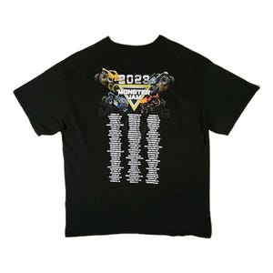 Puede incluir: Camiseta negra con un gr&aacute;fico colorido de monster trucks y el texto "2023 MONSTER JAM". Debajo del gr&aacute;fico hay listas de texto. La camiseta est&aacute; hecha de un material suave.