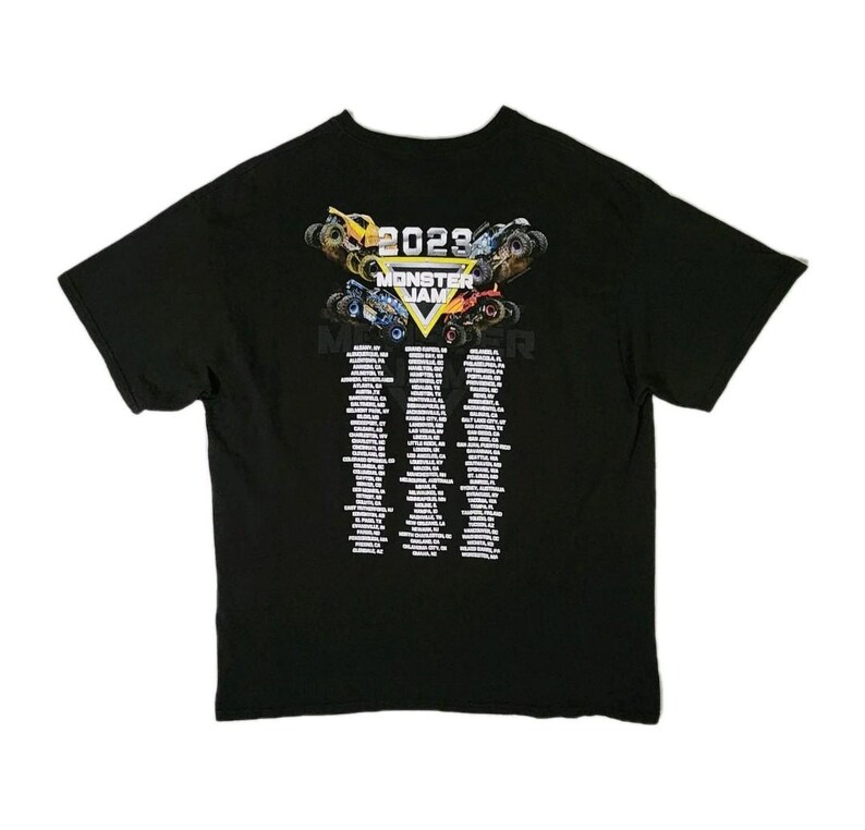 Puede incluir: Camiseta negra con un gr&aacute;fico colorido de monster trucks y el texto "2023 MONSTER JAM". Debajo del gr&aacute;fico hay listas de texto. La camiseta est&aacute; hecha de un material suave.