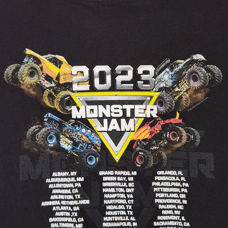 Puede incluir: Camiseta negra con un gr&aacute;fico de cuatro monster trucks en acci&oacute;n, el texto "2023 MONSTER JAM" en un dise&ntilde;o triangular y nombres de ciudades debajo. Un dise&ntilde;o din&aacute;mico para los fan&aacute;ticos de los eventos de monster trucks.
