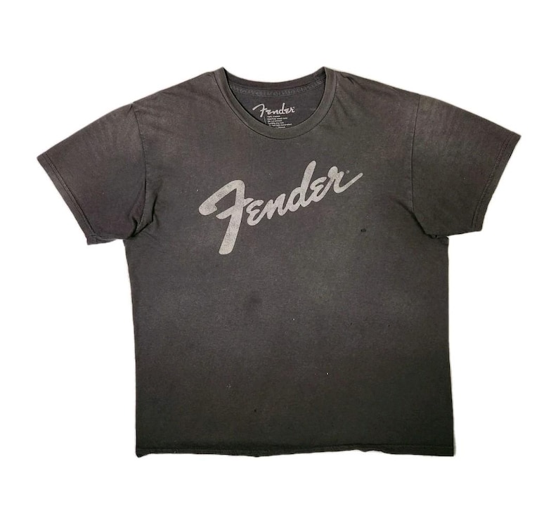 Puede incluir: Camiseta gris oscuro con el logotipo de Fender en escritura blanca en la parte delantera. La camiseta tiene cuello redondo y mangas cortas. El logotipo de Fender tambi&eacute;n est&aacute; impreso en la etiqueta.