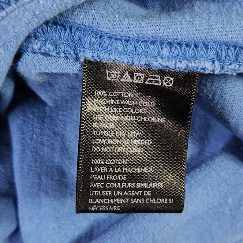 May include: A close-up of a blue fabric with a black tag. The tag features white text detailing care instructions: machine wash cold, tumble dry low, and low iron. The fabric is 100% cotton.
