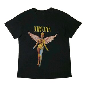 Peut inclure: T-shirt noir avec le nom du groupe "NIRVANA" en lettres dorées au-dessus d'un ange en mosaïque aux ailes déployées. Le corps de l'ange est un mélange de jaune, de bleu et de rouge.
